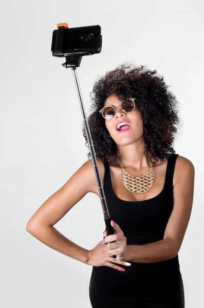 Hispanic model wearing sexy black dress, wild curly hair, golden necklace and sunglasses posing for selfie using selfiestick on white background — Stock Photo, Image