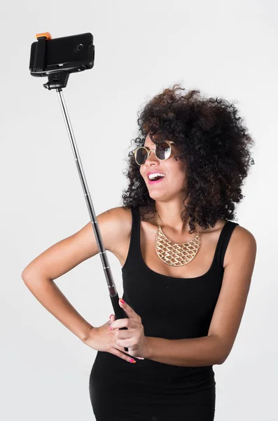 Hispanic model wearing sexy black dress, wild curly hair, golden necklace and sunglasses posing for selfie using selfiestick on white background — Stock Photo, Image