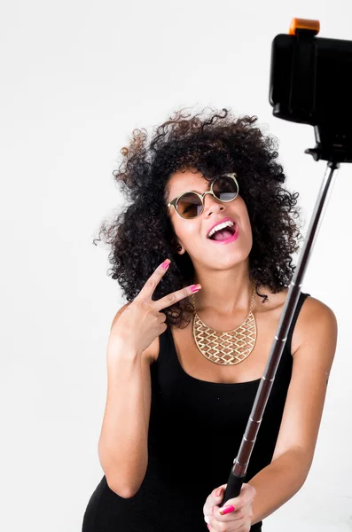 Hispanic model wearing sexy black dress, wild curly hair, golden necklace and sunglasses posing for selfie using selfiestick on white background — Stock Photo, Image