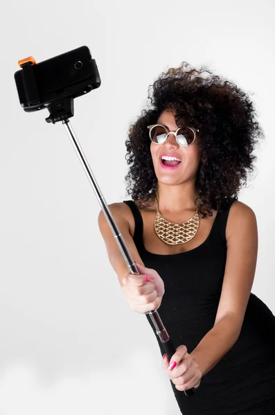 Hispanic model wearing sexy black dress, wild curly hair, golden necklace and sunglasses posing for selfie using selfiestick on white background — Stock Photo, Image