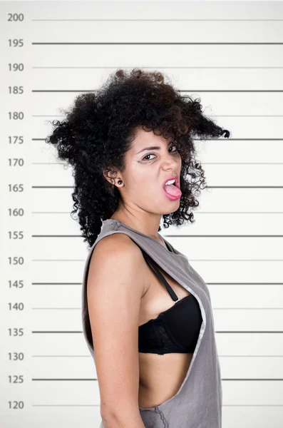 Modèle brune hispanique avec des cheveux salissants portant chemise sans manches grise regardant malheureux dans la caméra montrant la langue sous l'angle du profil, concept mugshot — Photo