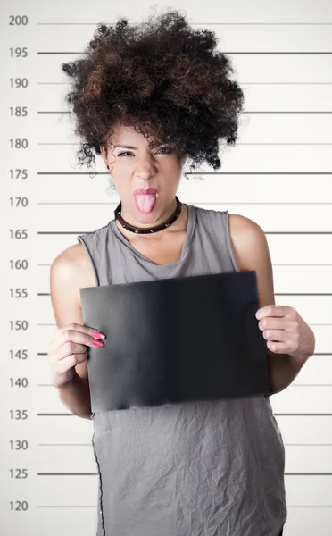 Hispanic brunette rebel model met afro zoals haar het dragen van grijze mouwloos shirt bedrijf leeg bord als poseren voor mugshot concept en tonen tong aan camera — Stockfoto