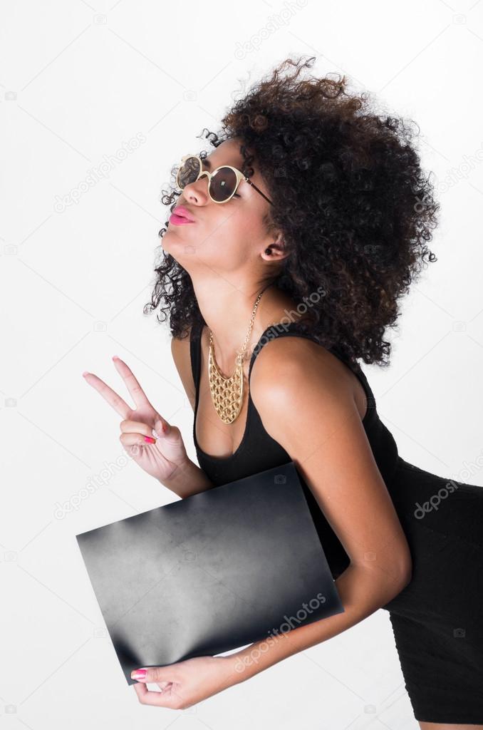 Hispanic model wearing black sexy dress and sunglasses holding blank board leaning forward making peace sign, shot from profile angle