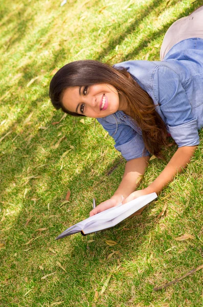 Brünettes Model, das glücklich im Gras liegt und ein Buch liest, aufgenommen aus dem Profilwinkel — Stockfoto