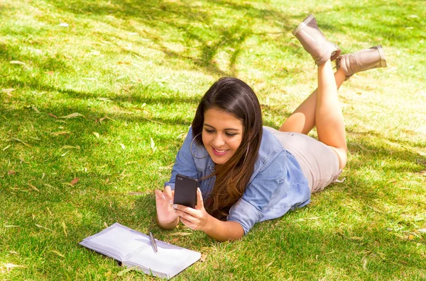 Modello bruna sdraiato su erba, piedi in aria attraversato giocare con il suo telefono sorridente — Foto Stock