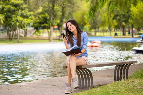 Brunette model wearing denim shirt and white shorts relaxing in park environment, sitting on bench next to lake looking at phone — 图库照片