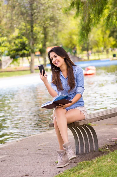 Brunette model wearing denim shirt and white shorts relaxing in park environment, sitting on bench next to lake looking at phone — Stock Photo, Image