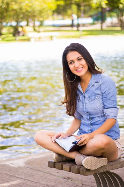 Modello bruna con camicia di jeans e pantaloncini bianchi che si rilassano nell'ambiente del parco, seduto sulla panchina accanto al lago sorridente alla macchina fotografica — Foto Stock