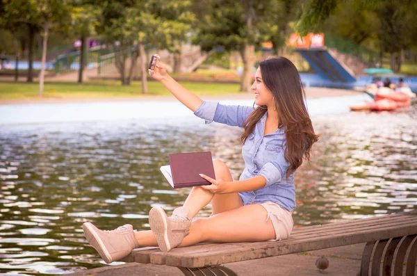 Brunette model wearing denim shirt and white shorts relaxing in park environment, sitting on bench next to lake taking selfie with mobile — 图库照片