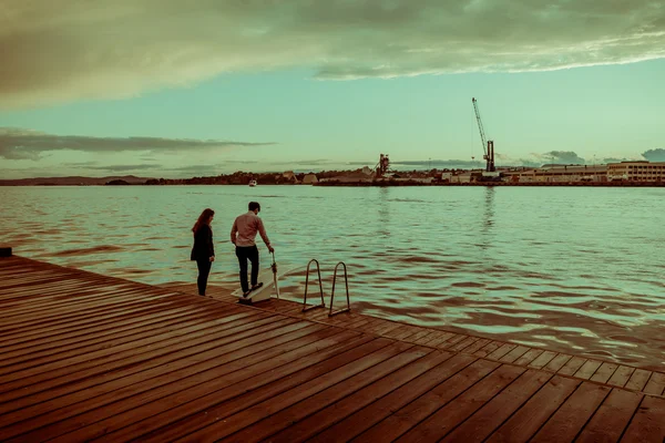 OSLO, NORVEGIA - 8 LUGLIO 2015: Due persone in piedi in acqua sul molo di legno di fronte al trampolino durante il tramonto, gru portuale distanza visibile — Foto Stock