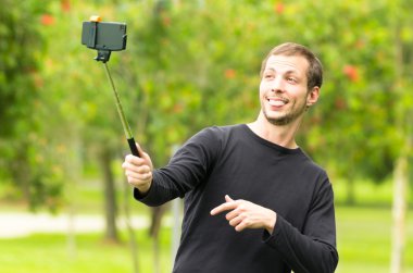 Hispanic man posing with selfie stick in park environment taking a photo of himself smiling