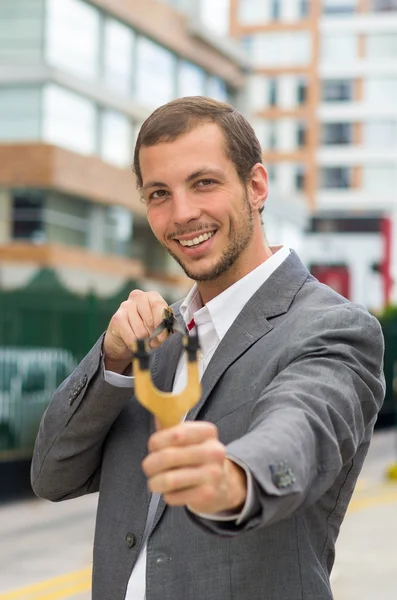 Handsome buisness man concentrated aiming a slingshot at urban city background — Zdjęcie stockowe