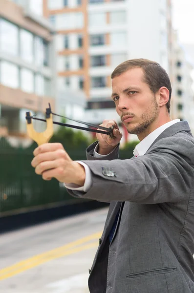 Handsome buisness man concentrated aiming a slingshot at urban city background — Zdjęcie stockowe