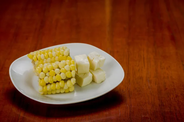 Assiette blanche avec la moitié de l'épi de maïs cuit couché à côté de morceaux de fromage carré frais stables — Photo