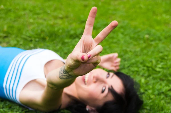 Headshot Brunetka zbliżenie poziomy leżąc na trawie z patrząc na kamery uśmiechając się i robi znak pokoju — Zdjęcie stockowe