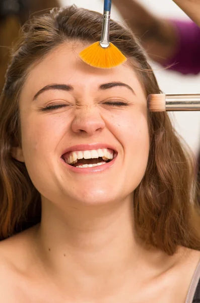 Closeup headshot brunette model facing camera laughing while getting makeup done by professional stylist using orange brush — 图库照片