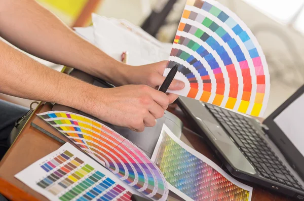 Manos masculinas sosteniendo la paleta de pantones, mapa de colores desplegado en frente de la computadora portátil en el escritorio de trabajo — Foto de Stock