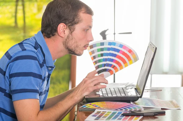 Mann arbeitet am Laptop, während er die Pantone-Palette hochhält, Farbkarte aus dem Profilwinkel — Stockfoto