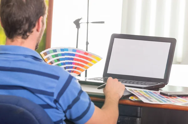 Mann arbeitet am Laptop, während er die Pantone-Palette hochhält, Farbkarte von hinten — Stockfoto