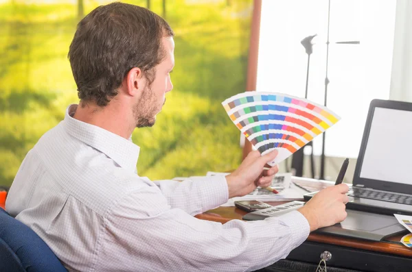 Mann arbeitet am Laptop, während er die Pantone-Palette hochhält, Farbkarte aus dem Profilwinkel — Stockfoto