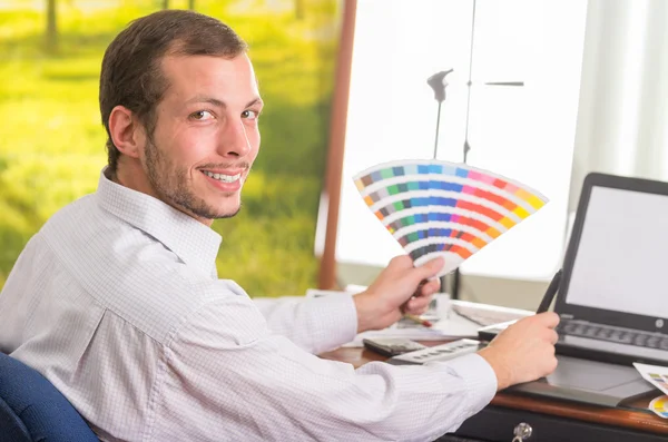 Hombre sonriendo a la cámara y trabajando en el ordenador portátil mientras sostiene la paleta pantone, colormap desde el ángulo del perfil —  Fotos de Stock