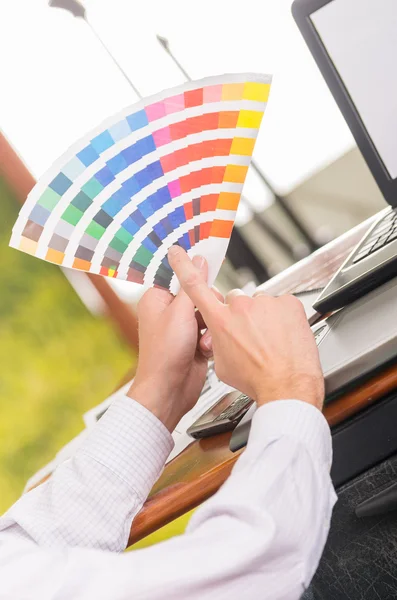 Manos masculinas sosteniendo la paleta de pantones, mapa de colores desplegado en frente de la computadora portátil en el escritorio de trabajo — Foto de Stock