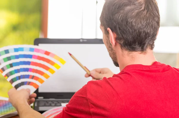 Man working on laptop while holding up pantone palette, colormap from behind angle — Stockfoto