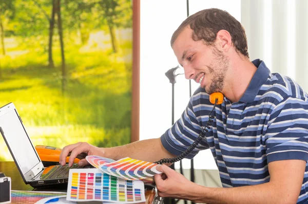 Man wearing blue white striped t-shirt sitting by work desk using phone and looking at pantone palette, colormap — Zdjęcie stockowe