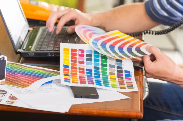 Mãos masculinas segurando paleta pantone, colormap espalhados na frente do laptop na mesa de trabalho — Fotografia de Stock
