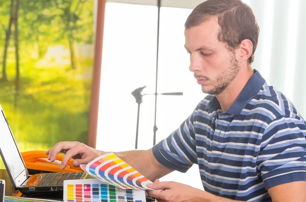 Man wearing blue white striped t-shirt sitting by work desk using phone and looking at pantone palette, colormap — Stock Photo, Image
