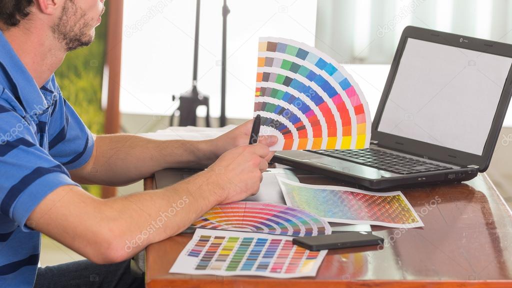 Male hands holding up pantone palette, colormap spread out in front of laptop on working desk