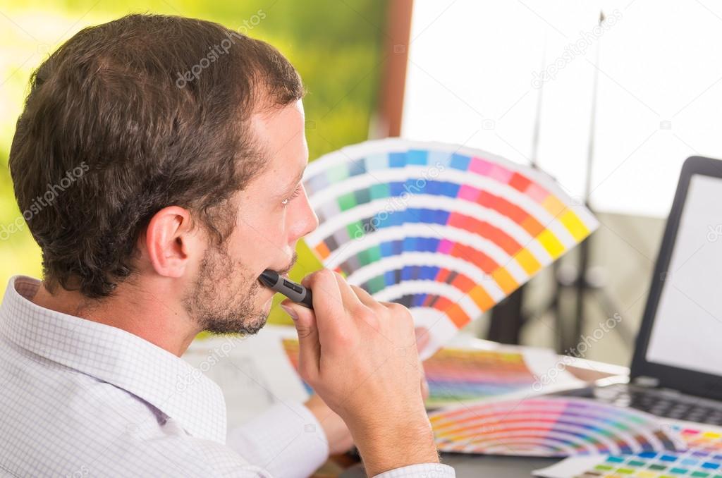 Man working on laptop while holding up pantone palette, colormap from behind angle