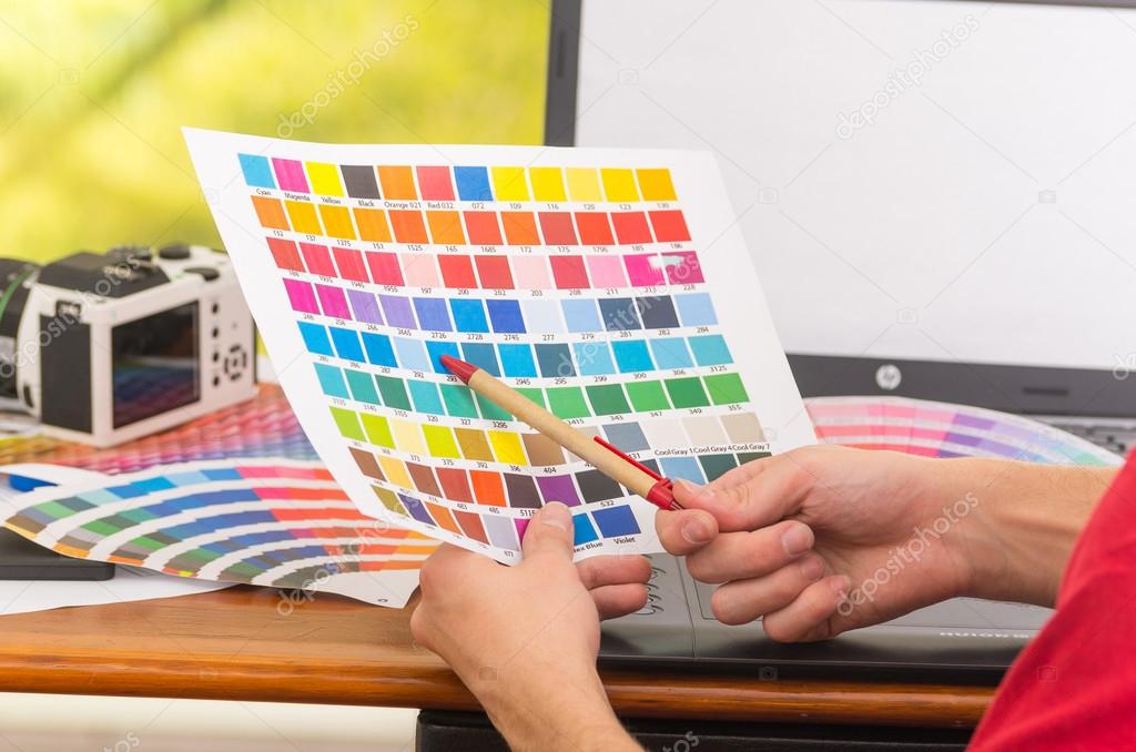 Male hands holding up pantone palette, colormap spread out in front of laptop on working desk