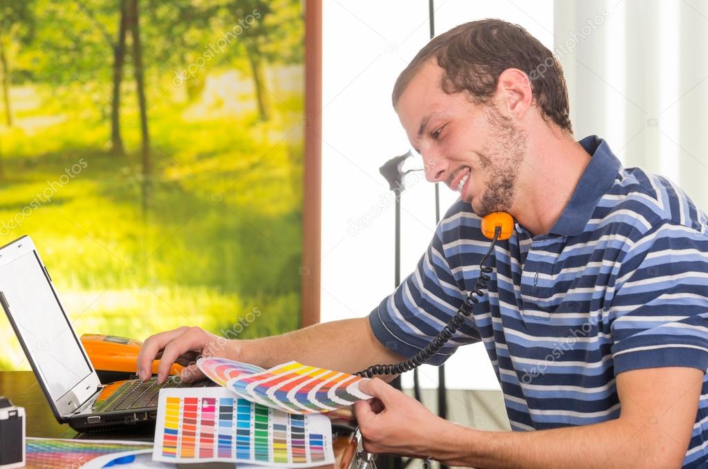 Man wearing blue white striped t-shirt sitting by work desk using phone and looking at pantone palette, colormap