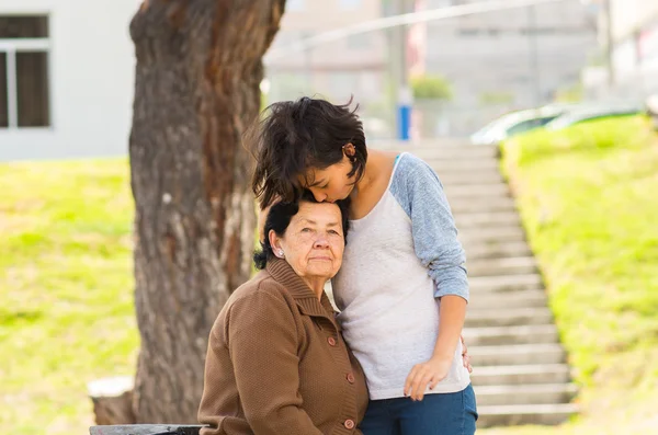 Young girl standing behind grandmother hugging and embracing in outdoors environment — Stock Photo, Image