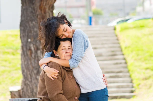 Young girl standing behind grandmother hugging and embracing in outdoors environment — Stock Photo, Image