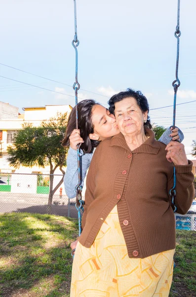 Молодая девушка, сидящая на качелях и целующая бабушку в щеку, с радостью позирует перед камерой — стоковое фото