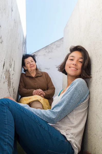 Schöne hispanische Großmutter Enkelin in gelbem Rock und brauner Jacke sitzt, während sie in die Kamera schaut — Stockfoto