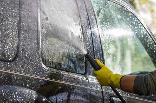 Arm with yellow glove holding high pressure water cleaner and using it on car door windows — 图库照片