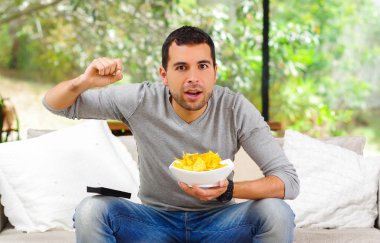 Hispanic male wearing light blue sweater plus denim jeans sitting in white sofa holding bowl of potato chips and remote control watching tv enthusiastically