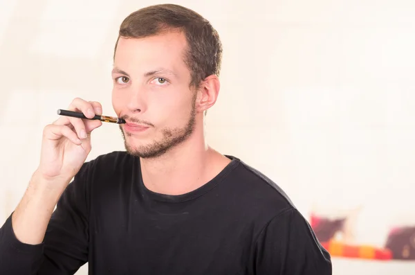 Hombre hispano con suéter oscuro disfrutando de un cigarrillo electrónico mientras posa para la cámara —  Fotos de Stock