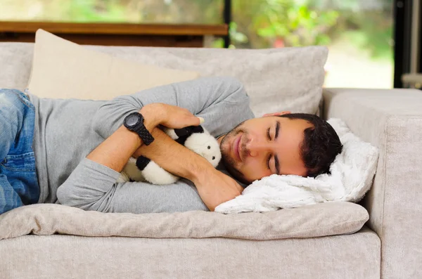Hispanic male wearing blue sweater and jeans lying on white sofa with stuffed animal between arms sleeping — 스톡 사진