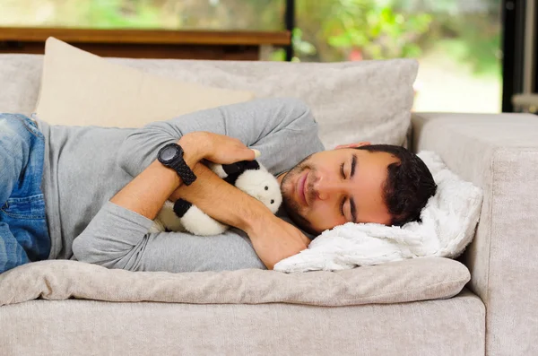 Spanischer Mann mit blauem Pullover und Jeans auf weißem Sofa liegend mit Stofftier zwischen den Armen schlafend — Stockfoto