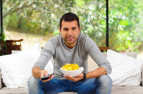 Hombre hispano vistiendo suéter azul claro más vaqueros sentados en un sofá blanco sosteniendo tazón de papas fritas y control remoto viendo la televisión con entusiasmo —  Fotos de Stock