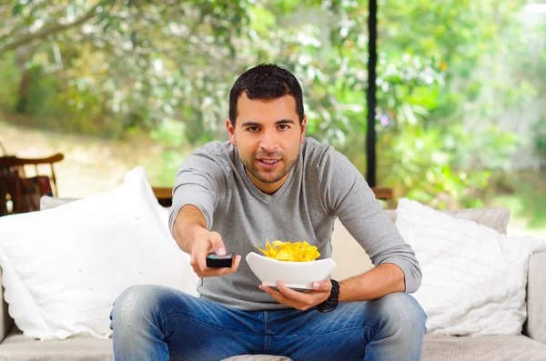 Homem hispânico vestindo camisola azul claro mais jeans jeans sentados no sofá branco segurando tigela de batatas fritas e controle remoto assistindo tv entusiasticamente — Fotografia de Stock