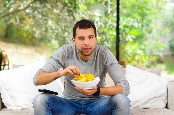 Hispanic male wearing light blue sweater plus denim jeans sitting in white sofa holding bowl of potato chips and remote control watching tv enthusiastically — Zdjęcie stockowe