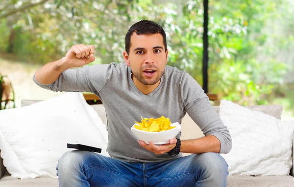 Hispanic male wearing light blue sweater plus denim jeans sitting in white sofa holding bowl of potato chips and remote control watching tv enthusiastically — Stock Fotó