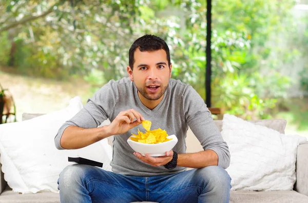 Hispanic male wearing light blue sweater plus denim jeans sitting in white sofa holding bowl of potato chips and remote control watching tv enthusiastically — Zdjęcie stockowe