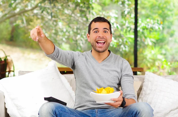 Hispanic male wearing light blue sweater plus denim jeans sitting in white sofa holding bowl of potato chips and remote control watching tv enthusiastically — Zdjęcie stockowe