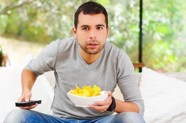 Hispanique mâle portant un pull bleu clair plus jeans en denim assis dans un canapé blanc tenant bol de croustilles de pomme de terre et télécommande regarder la télévision avec enthousiasme — Photo